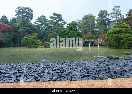 A tranquil landscape featuring a calm green pond in a Japanese garden setting, reflecting the sky and trees, with a well-defined stone border and a pr Stock Photo