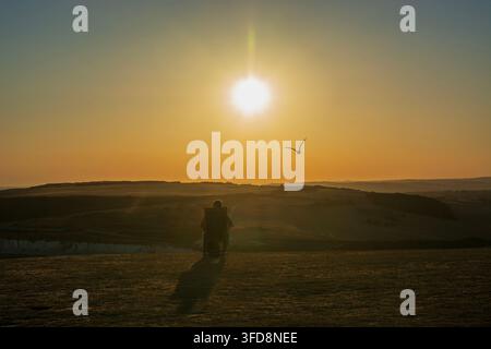 A lone man sitting in chair  on Seven Sisters Cliff enjoying sunset with seagull flying over his head, East Sussex., UK Stock Photo