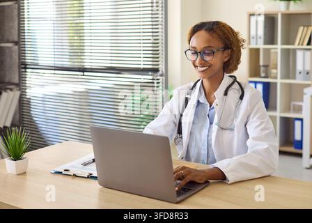 Smiling Female Doctor Using Laptop at Desk in Modern Medical Office Healthcare Stock Photo