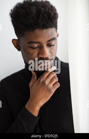 A young Black man with a stylish afro hairstyle gazes downward with a pensive expression, his hand resting on his chin Stock Photo