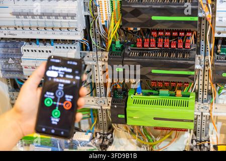 A hand holds a smartphone displaying an energy management app next to a complex electrical control system with many wires and modules Stock Photo
