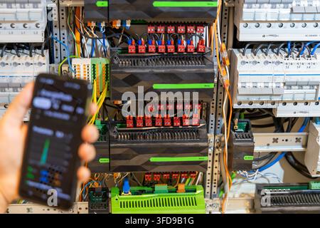 A hand holds a smartphone displaying a control interface next to a complex electrical panel with relays and wiring Stock Photo
