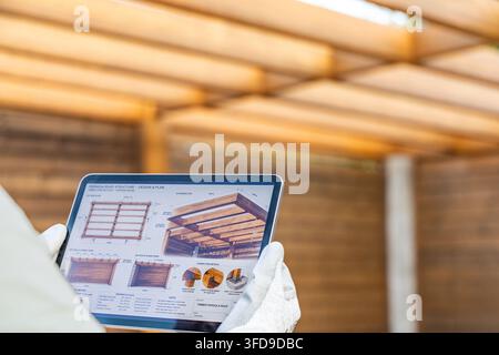 A person wearing gloves holds a tablet displaying detailed blueprints and 3D renderings of a wooden pergola structure Stock Photo
