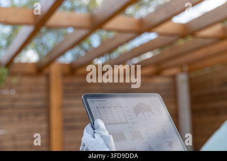 A gloved hand holds a tablet displaying architectural blueprints for a wooden structure, with the actual pergola visible in the background Stock Photo