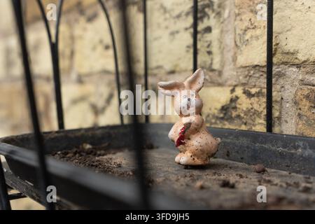 A small, weathered ceramic rabbit sits on a black metal garden shelf against a stone wall, functioning as a decorative outdoor ornament. Stock Photo