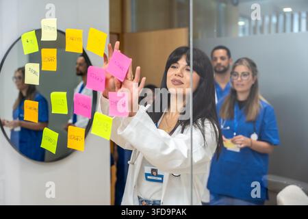 Medical doctor placing colorful sticky notes on a glass whiteboard while a diverse healthcare team collaborates in a modern clinic to brainstorm strat Stock Photo