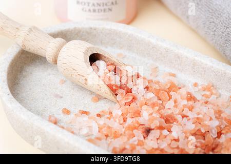 Pink Himalayan salt crystals spilling from a wooden scoop in a stone bowl with botanical skincare products. Closeup. Stock Photo