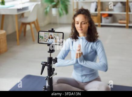 Woman sitting in lotus pose, calmly on mat with eyes closed hands in nanaste, recording online Stock Photo