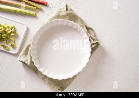 Empty white ceramic pie dish with chopped fresh rhubarb on bright kitchen background. Baking preparation concept with copy space. Top view, flat lay, above. Stock Photo