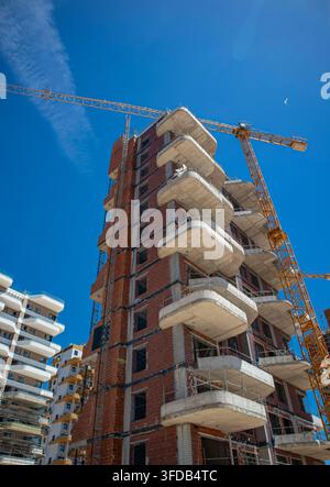 The view shows a tall residential building being built. A large yellow tower crane stands beside the structure. The sky is bright and blue. Work is on Stock Photo