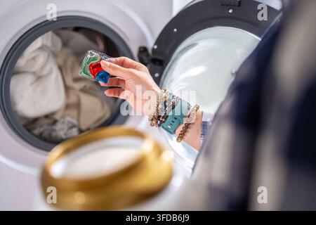 Hand loads colorful laundry detergent pod into open front-loading washing machine. Stock Photo