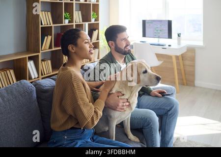 Happy couple with labrador dog relaxing on sofa and watching TV at home together Stock Photo