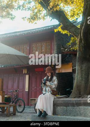 Woman reviewing camera photos under golden ginkgo tree, Lijiang Old Town, Yunnan, China Stock Photo