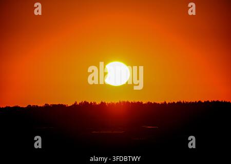 The crescent-shaped sun glows above a dark treeline during the partial solar eclipse of August 12, 2026, as the moon covers part of its disc. The deep Stock Photo