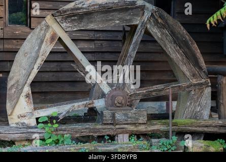 An old weathered wooden water wheel stands in front of a dark wooden wall. Stock Photo