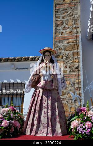 Virgin Mary stands serene, adorned in ornate, traditional robes. Floral arrangements frame her, adding grace to the scene. Stone wall and clear sky pr Stock Photo