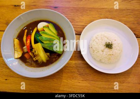 Delicious Japanese soup curry served in a bowl with various fresh vegetables including avocado and boiled egg, accompanied by a side of white rice on Stock Photo