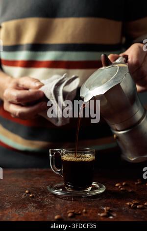 A man in a striped sweater is poured coffee from a geyser coffee maker into a glass cup. It's an authentic vintage Italian coffee maker, a 1950s and 1 Stock Photo