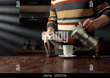 A man in a striped sweater is poured coffee from a geyser coffee maker into a white cup. It's an authentic vintage Italian coffee maker, a 1950s and 1 Stock Photo