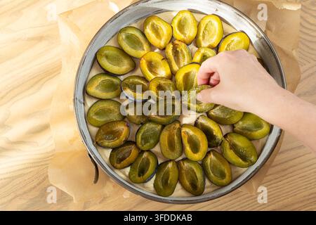 Woman hand placing fresh plum halves on raw dough in baking tin preparing plum pie Stock Photo