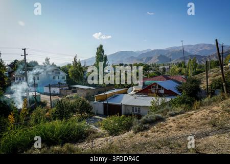 A rural Kyrgyz village is nestled on a sloping hillside with scattered houses, green trees, and utility poles, while smoke rises from a few properties Stock Photo
