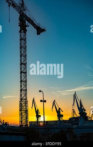 Shipyard cranes silhouetted against sunset sky. Industrial shipyard cranes stand in silhouette against a vivid blue and golden sunset sky above the Stock Photo