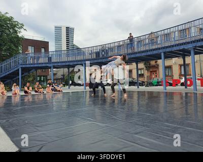 Three contemporary dancers perform an acrobatic lift in an outdoor urban plaza. Two dancers support a third dancer horizontally above their shoulders Stock Photo