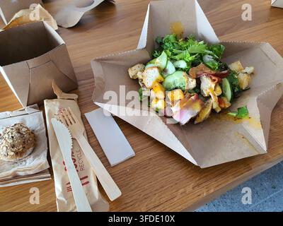 A top-down view of a fresh salad served in a recyclable cardboard takeout container on a wooden table. The colorful salad contains mixed greens, Stock Photo