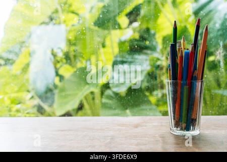 Colored pencils in a glass cup on a wooden table against a window with lush green foliage. High quality photo Stock Photo