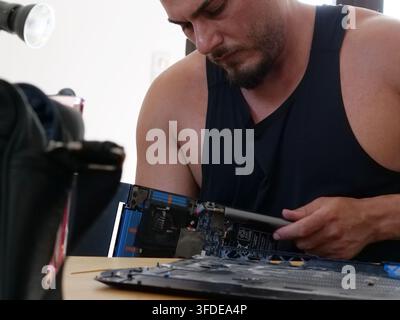 Close-up view of a bearded technician in a black tank top carefully using an electric precision screwdriver on an opened laptop motherboard. The focus Stock Photo