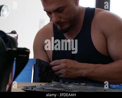 Focused front shot of a bearded technician in a black tank top working intently on an opened laptop resting on a wooden table. His hands are positione Stock Photo