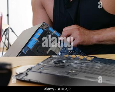 Close-up perspective of a muscular man's hands in a black tank top, working precisely on the interior of a laptop. His finger is near the fan assembly Stock Photo