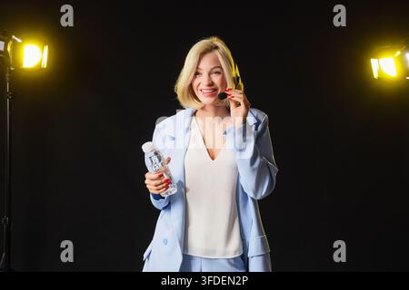 Customer support and remote communication, smiling woman with headset holding water bottle in professional studio setting with bright lighting Stock Photo