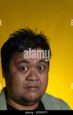An expressive close up portrait displays a young Asian male with wide open eyes and a shocked facial expression against a vivid yellow background. The Stock Photo