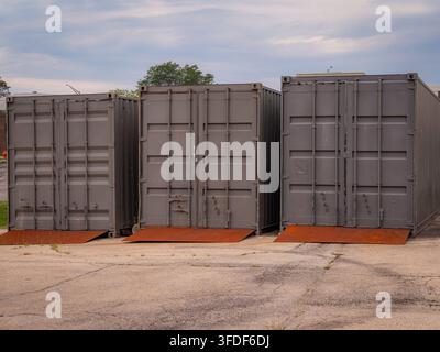 Three grey metal shipping containers with rusted loading ramps parked on an asphalt lot outdoors Stock Photo