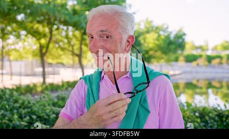 Senior man smiling holding eyeglasses enjoying outdoor park Stock Photo