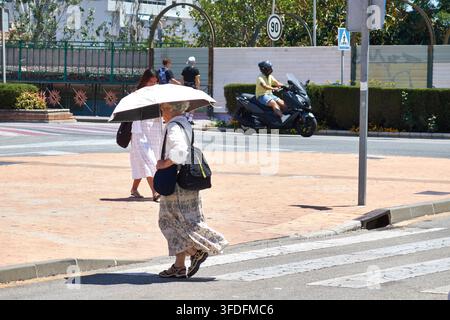 Elderly woman walking on a crosswalk with a sun umbrella to protect herself from the heat on a sunny day. Stock Photo