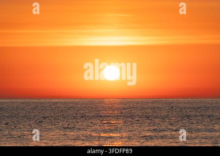 Bright glowing sun rising over calm ocean horizon with water reflections under orange sky Stock Photo