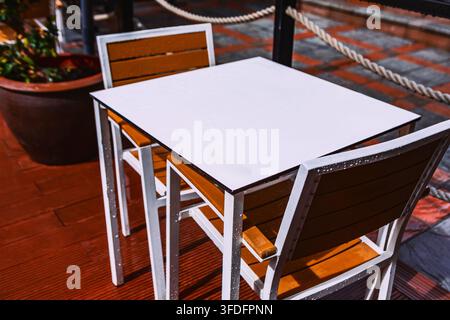 Outdoor cafe table with white square top and wooden slatted chairs stands on a deck. Rope railings and potted plants create a relaxed patio setting Stock Photo