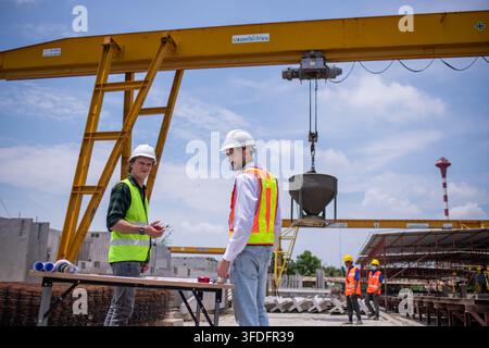 Engineers wearing safety helmets and reflective vests working construction site large overhead crane operation ensure efficient secure handling of hea Stock Photo