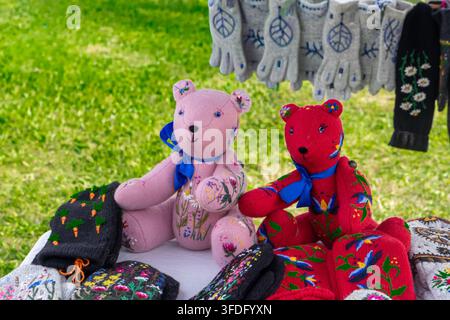 Handmade pink and red teddy bears with floral embroidery displayed on table at outdoor craft fair. Soft fabric bear toys with blue ribbon bows sitting Stock Photo