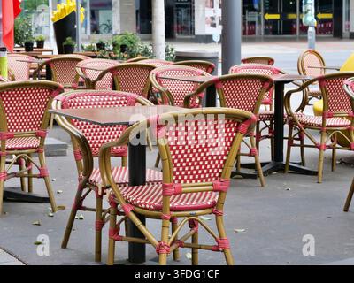 Wide view of an empty outdoor street cafe seating area featuring classic Parisian-style woven red and white chairs and small square dark tables. Set o Stock Photo