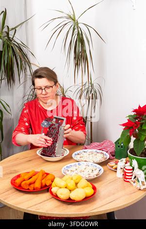 A woman grates beets for the Shuba salad on a Christmas table. Various ingredients are laid out, including herring, potatoes, eggs, and carrots, creat Stock Photo