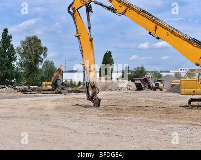 Wide angle shot of a large yellow hydraulic demolition excavator arm positioned over ground on a vast industrial clearing site. Features distant const Stock Photo