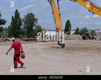 Worker wearing red safety clothing walking across a dusty industrial construction and demolition ground while holding a power tool. Massive yellow hyd Stock Photo