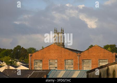 View across red brick industrial rooftops in Radcliffe towards the historic tower of St Thomas and St John's Church under a dramatic cloudy sky. Stock Photo