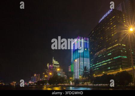 A panoramic nighttime waterfront cityscape features illuminated high-rise buildings, colorful architectural lighting, bright streetlights, and reflections on the water. Stock Photo