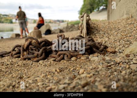 A heavy rusted metal chain lies on a rocky shoreline beside a stone wall, with two blurred people and calm water in the background. Shallow depth of f Stock Photo