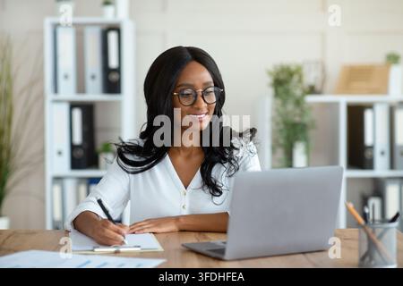 Cheerful black business lady working on laptop, taking notes during online work Stock Photo