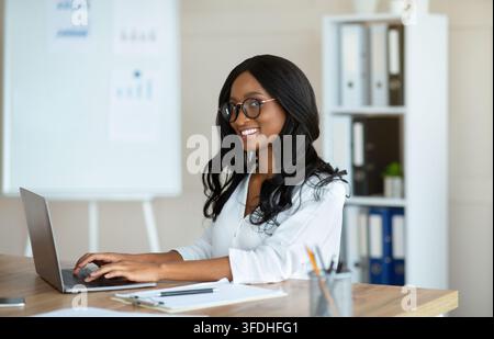 Happy young black businesswoman in glasses sitting at desk with open laptop Stock Photo
